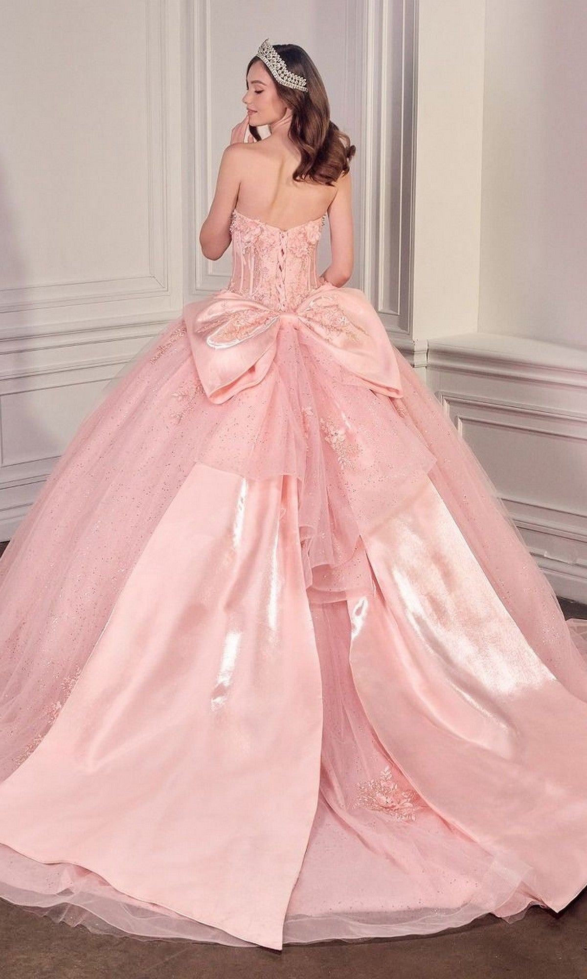 A woman faces away in a decorated room, wearing the Quinceanera Dress 15726 By Ladivine—a pink glitter tulle ball gown with a large bow—topped with a sparkling tiara.
